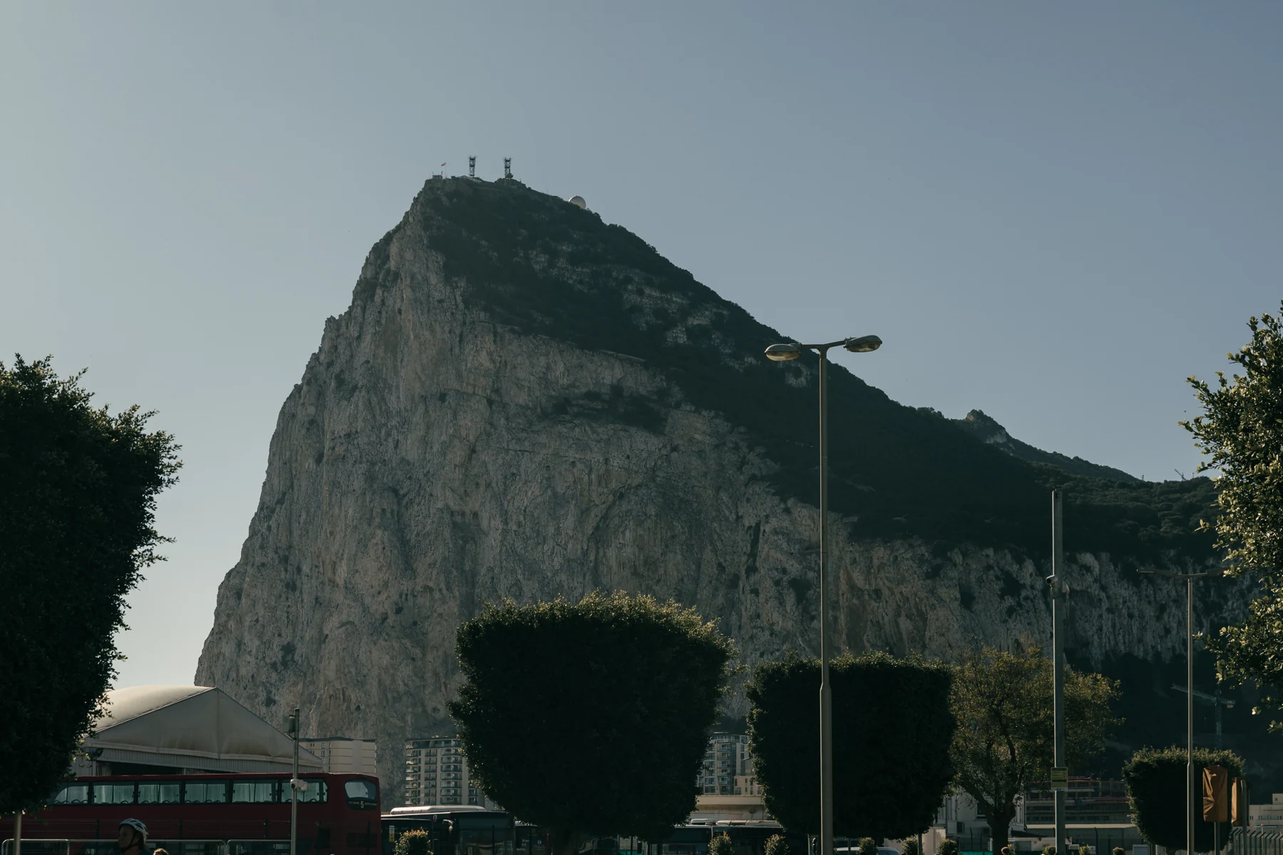 The north face of the Rock of Gibraltar rises above trimmed trees, street lamps and a red double-decker bus on a clear morning.