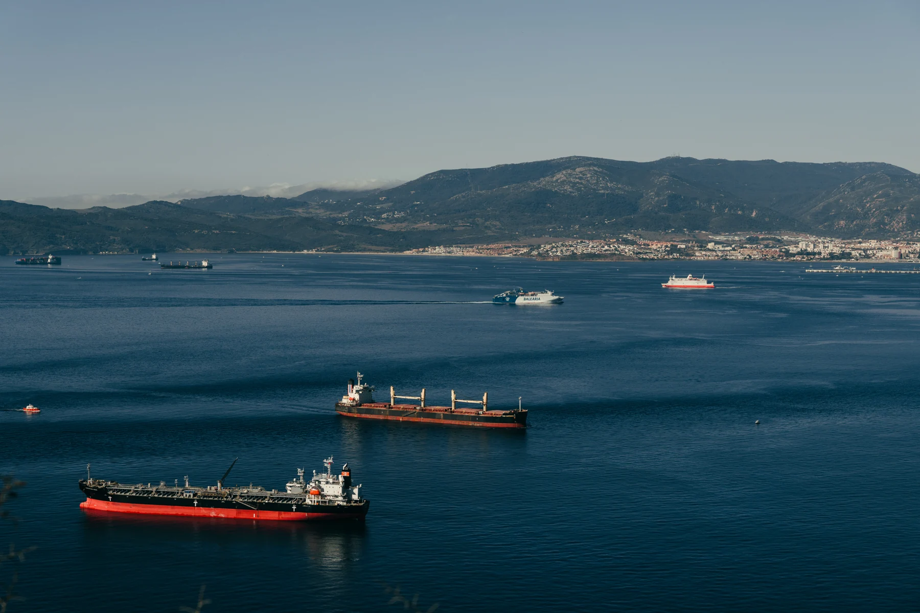 Two red-hulled cargo ships at anchor in the calm Bay of Gibraltar, with the city of Algeciras and the hills of Andalusia on the far shore.