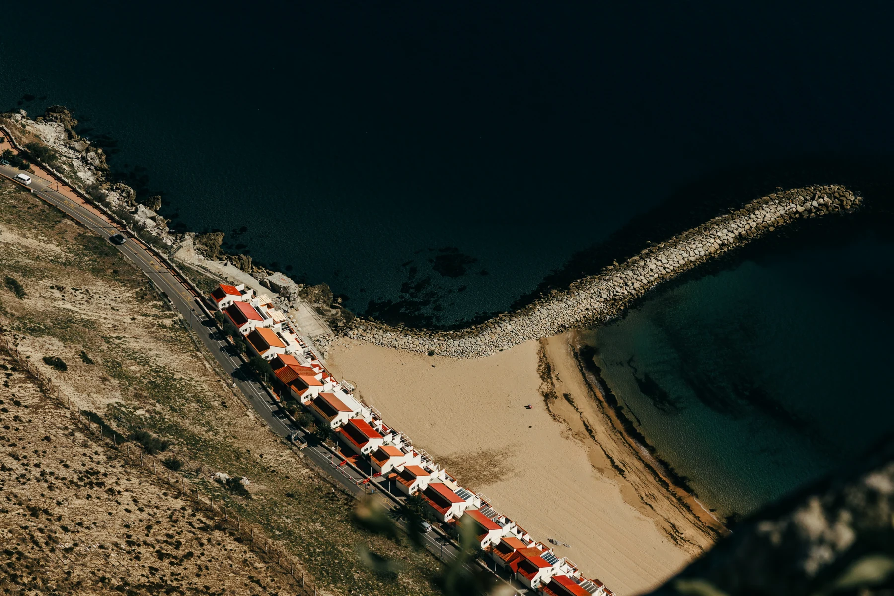 Looking straight down from the Rock onto a sandy beach, a curved stone breakwater and a row of houses on Gibraltar's east coast.