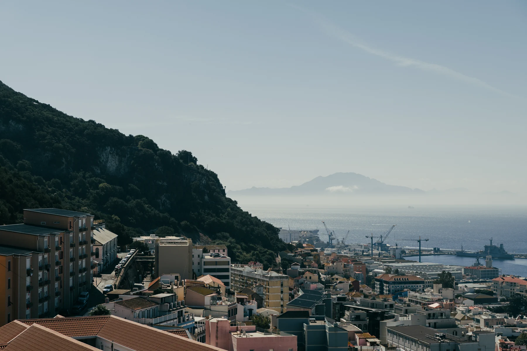 Terracotta rooftops of Gibraltar's town beneath the Rock, with Jebel Musa in Morocco visible across the hazy Strait.