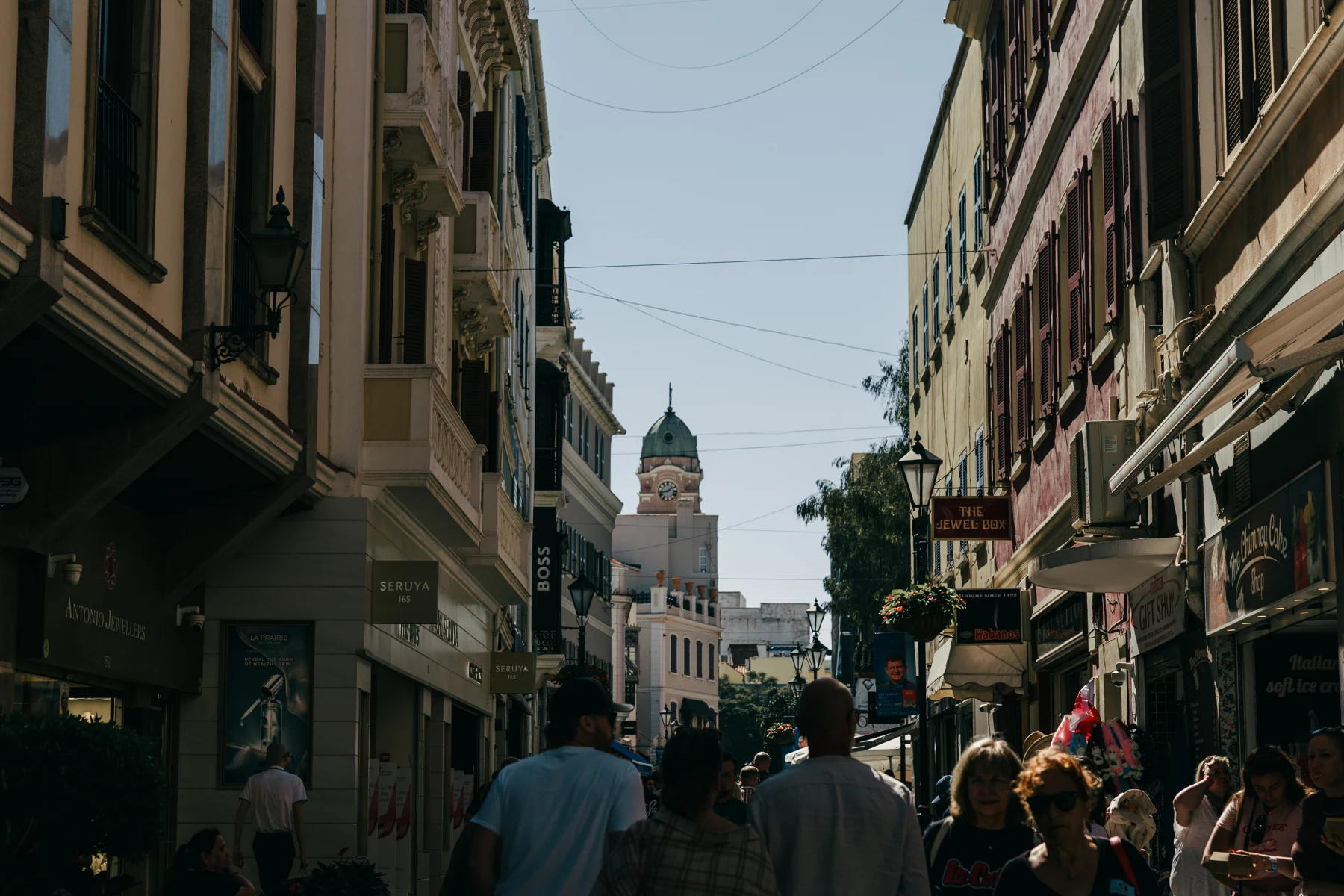 Shoppers and tourists walk along a narrow shopping street in Gibraltar town toward a domed tower.